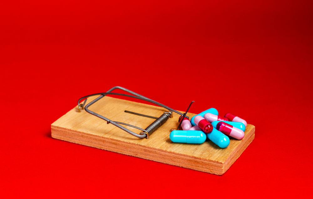 A wooden mousetrap against a red background. Pills are the bait.