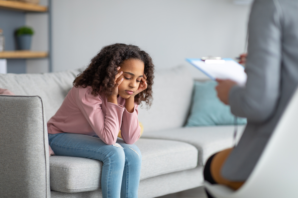 Unhappy girl listening to psychologist at meeting. Doctor consulting stressed teen at counselling therapy session. Little patient sitting on couch at office or home, thinking about her problems