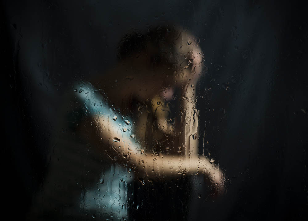 A girl looks sad, blurry through a rainy pane of glass, dark