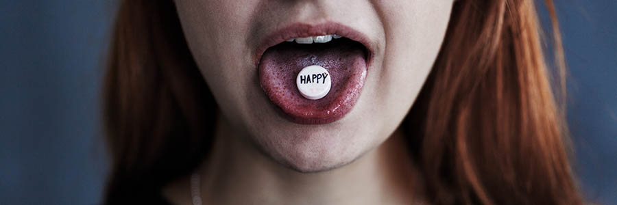 Panorama with close-up of girl's mouth with white pill on the tongue. Pill has text reading "HAPPY" on it.