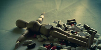 An artist's posable mannequin lies flat amongst a pile of pills, dark lighting