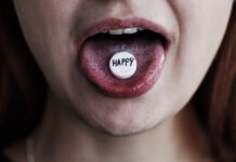 In Advancing Psychedelics, Australian Drug Regulator Ignored the Report of Its Own Researchers close-up of girl's mouth with white pill on the tongue. Pill has text reading "HAPPY" on it.