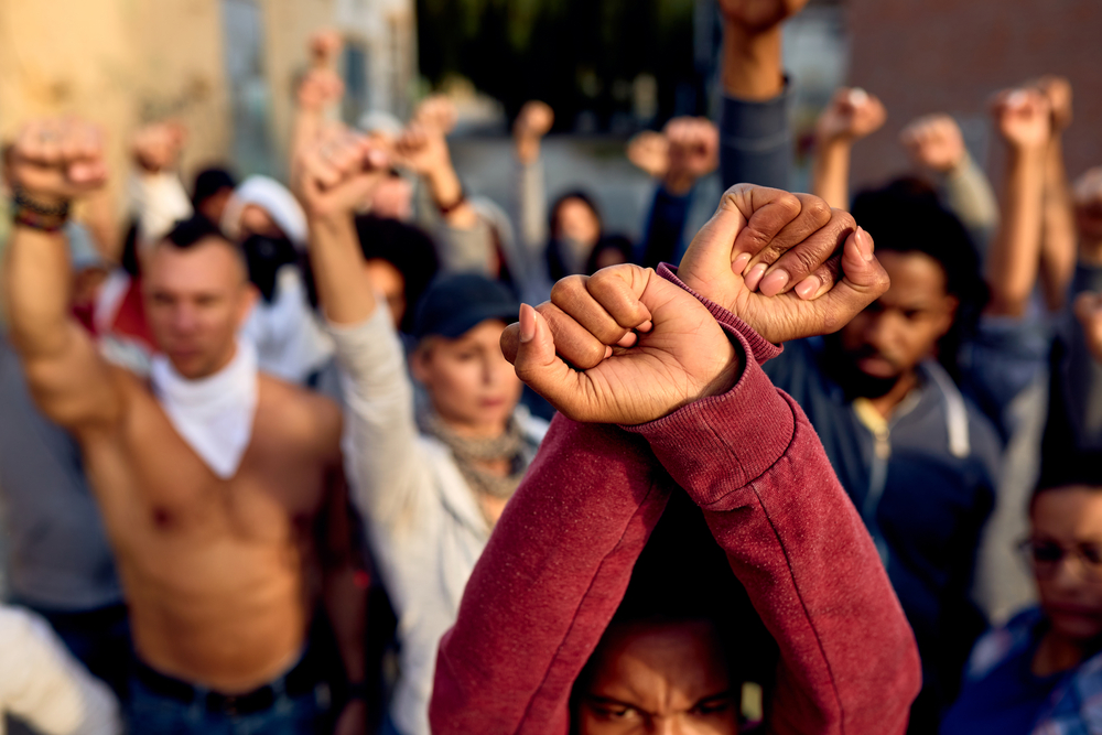 Close-up of black woman with clenched fists above her head protesting with group of people on the streets.