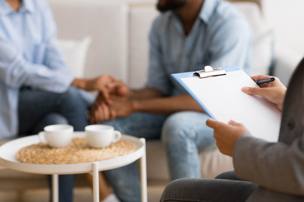 A black couple holds hands out of focus in background. In foreground, a therapist's hands on a clipboard taking notes.