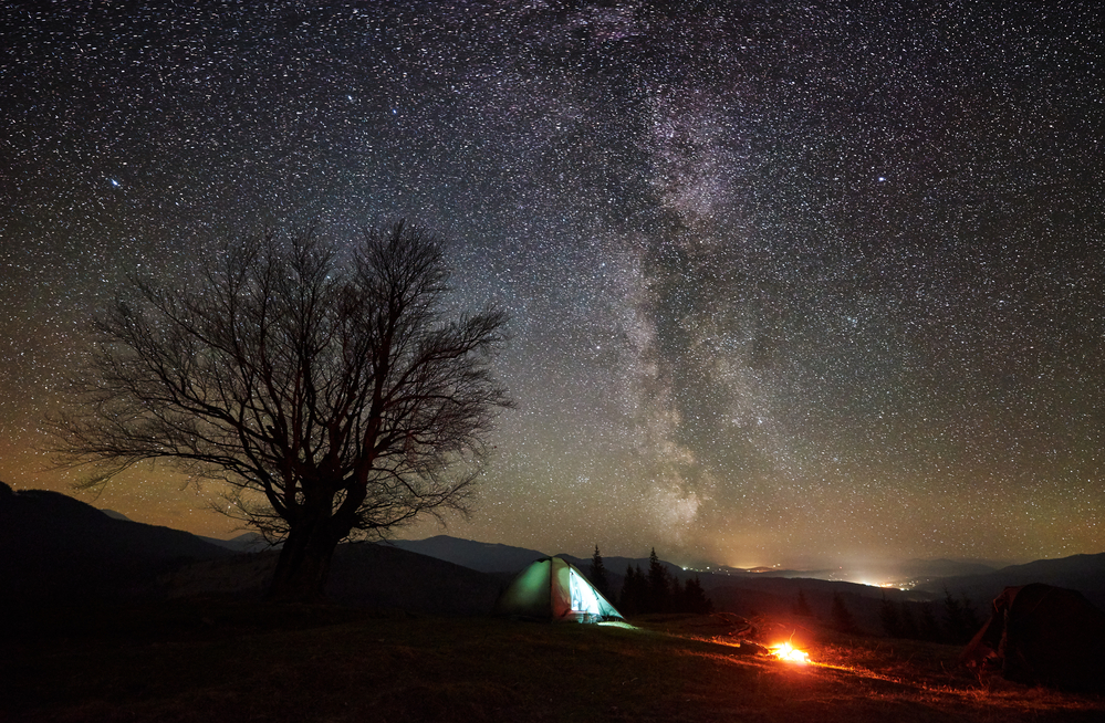 Campfire burning near tourist illuminated tent. Night camping in mountains under starry sky and Milky way. Silhouette of big tree and distant hills on background. Tourism, outdoor activity concept