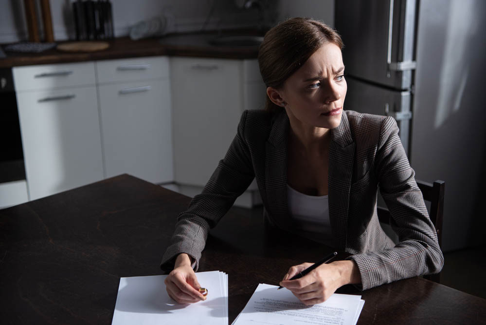 A woman looks to the side in distress, while holding a wedding band and a pen to sign divorce documents