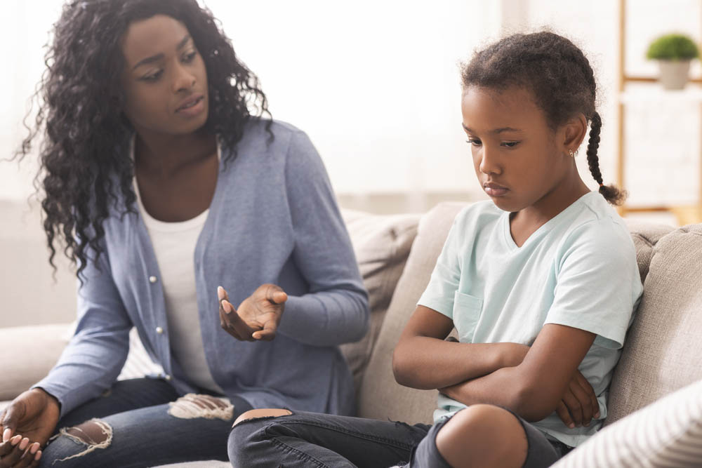 A Black mother and daughter sit on the couch. Mother is explaining something to a sullen-looking girl.