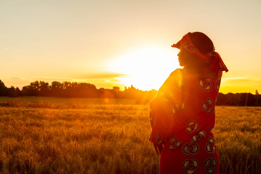 African woman in traditional clothes standing, looking, hand to eyes, in field of barley or wheat crops at sunset or sunrise