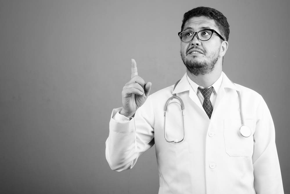 Studio shot of bearded Persian man doctor against gray background in black and white