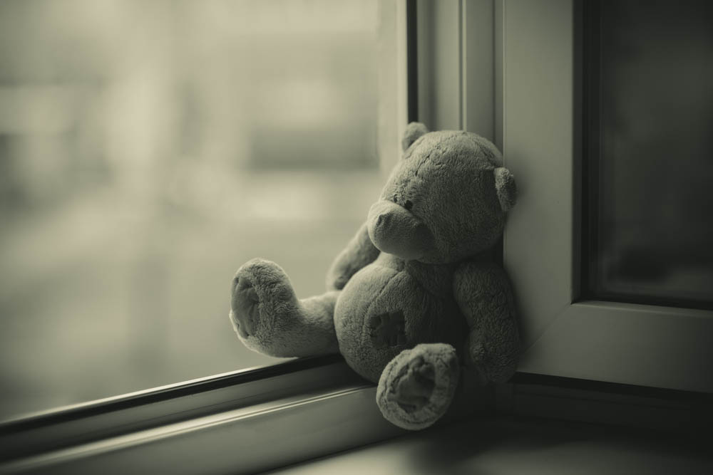 A sepia-tone photograph of a lonely teddy bear small against a window.