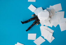 The Co-Opting of the Peer Movement in Mental Health Miniature photography. A man's legs are visible under a pile of paperwork on a blue background.