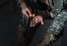 Risks of Prescribing Psychiatric Drugs to Veterans A hand holding pills from a pill bottle. Legs in military camo are visible