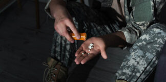 Risks of Prescribing Psychiatric Drugs to Veterans A hand holding pills from a pill bottle. Legs in military camo are visible