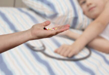 For-Profit Healthcare Is a Predator; Its Main Prey Is Our Young A hand holds out pills. A child is out of focus in the background.
