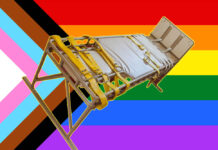 A hospital bed with restraints. The background is a pride flag with all colors.