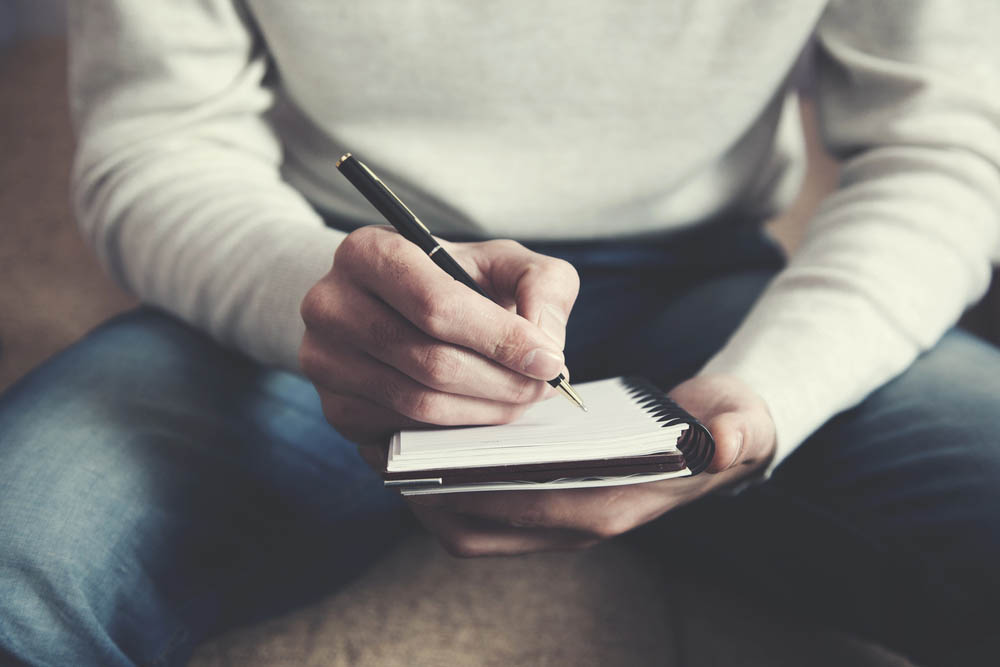 A man is sitting on a sofa and writing notes in a notebook
