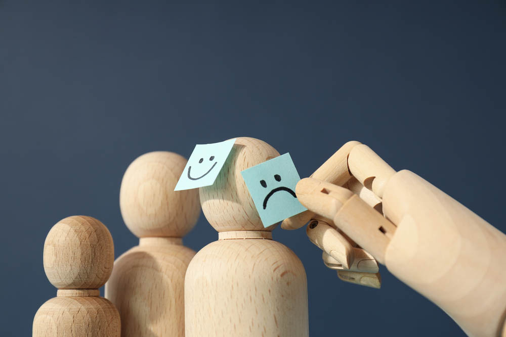 A wooden hand touches post it notes of smiling and sad faces on wooden figures