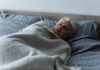 A young blonde woman looks sad in bed with the covers up to her neck