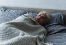 A young blonde woman looks sad in bed with the covers up to her neck
