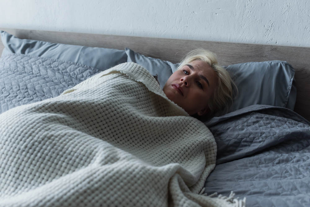 A blonde woman looks sad in bed with the covers up to her neck