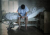 Man in hospital gown sits on hospital bed with head bowed in dark room