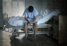 Man in hospital gown sits on hospital bed with head bowed in dark room
