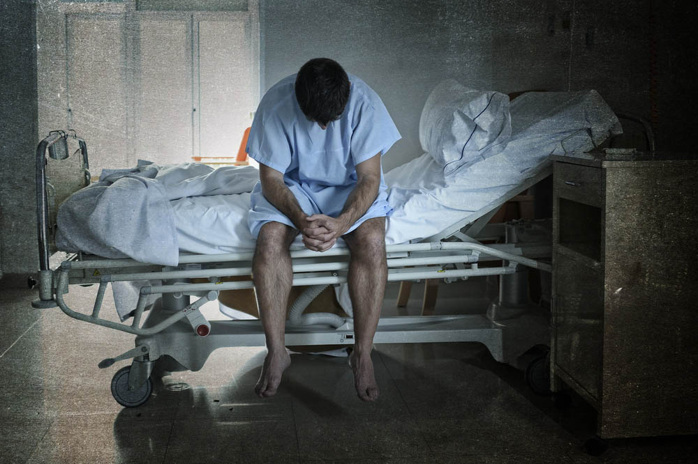 Man in hospital gown sits on hospital bed with head bowed in dark room