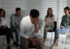 Unhappy man and group of people behind his back indoors. Therapy session