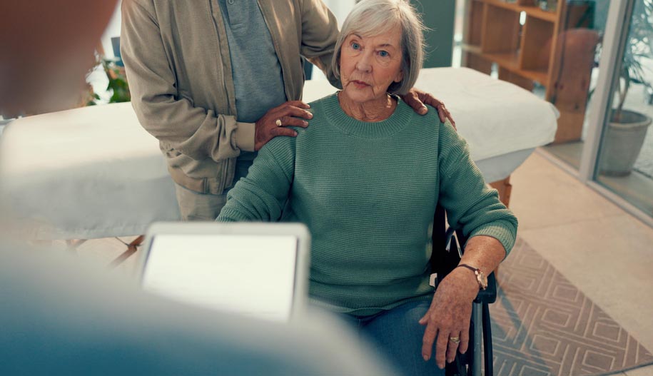 An elderly woman in a wheelchair tells a doctor (out of focus) about her symptoms