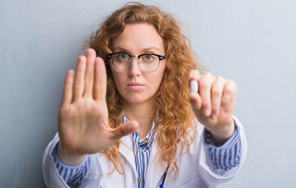 white professional woman holds a pill in one hand and makes a "stop" gesture with the other