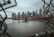 Addressing the Twofold Mental Health Crisis in New York City View of New York City skyline through a chain-link fence, featuring the Brooklyn Bridge and skyscrapers under a cloudy sky.