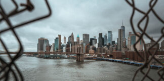Addressing the Twofold Mental Health Crisis in New York City View of New York City skyline through a chain-link fence, featuring the Brooklyn Bridge and skyscrapers under a cloudy sky.