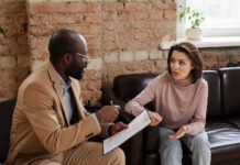 Beyond Medication: What England’s Experience Can Teach Us About Psychosis Care African-American psychologist in eyeglasses sitting with clipboard and working with problems of patient in loft office