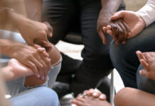 The Mental Health Crisis We’re Not Naming Close up diverse people sitting on chairs in circle, holding hands at group therapy
