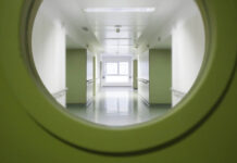 The Person They Never Asked About Looking through a round window in a green door into a white hospital corridor