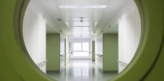 The Person They Never Asked About Looking through a round window in a green door into a white hospital corridor
