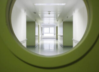 The Person They Never Asked About Looking through a round window in a green door into a white hospital corridor